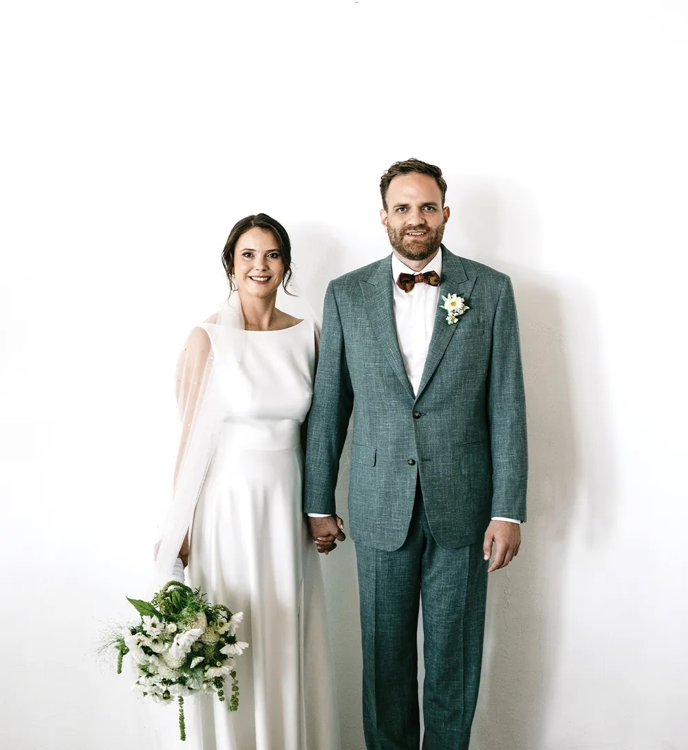 The bride and groom standing hand in hand in front of a white wall